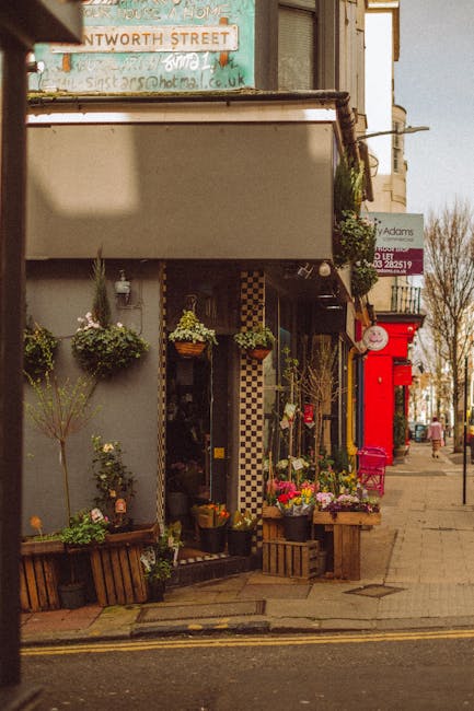 The exterior of a flower shop on Hoe Street Walthamstow E17 displays an inviting arrangement of floral displays in wooden crates and hanging baskets, featuring vibrant pink, white, and green blooms. F