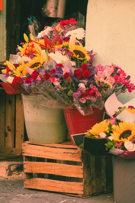 A collection of vibrant floral bouquets arranged in various containers, including a large beige ceramic pot, a red plastic bucket, and a smaller black box, placed on a wooden crate outdoors. The bouqu